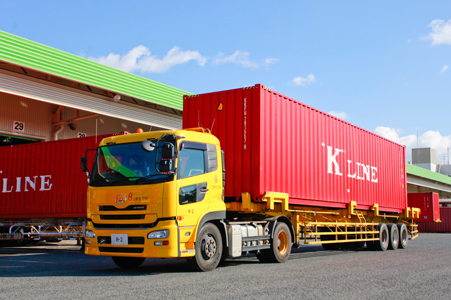 A photo of a yellow truck carrying a red KLINE container, parked in front of a warehouse.
